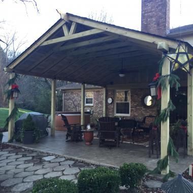 A covered patio with a gabled wooden roof, stone floor, outdoor furniture, and holiday greenery attached to pillars.
