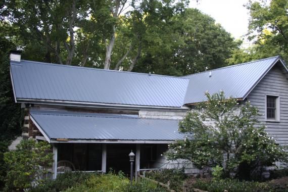 A rustic, multi-section cabin with gray siding and a light gray metal roof, surrounded by trees and lush greenery.