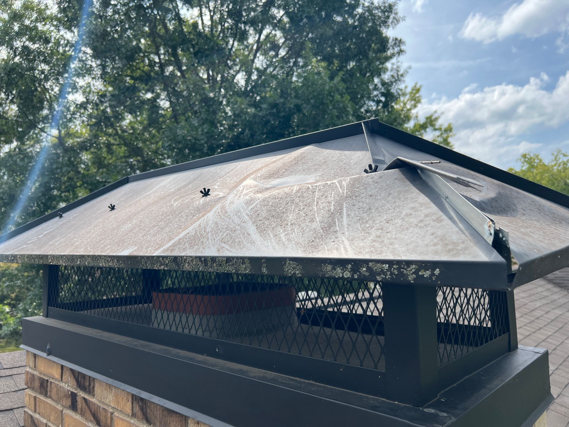 A damaged metal chimney cap with a bent corner section, sitting atop a brick chimney on a roof against a blue sky.