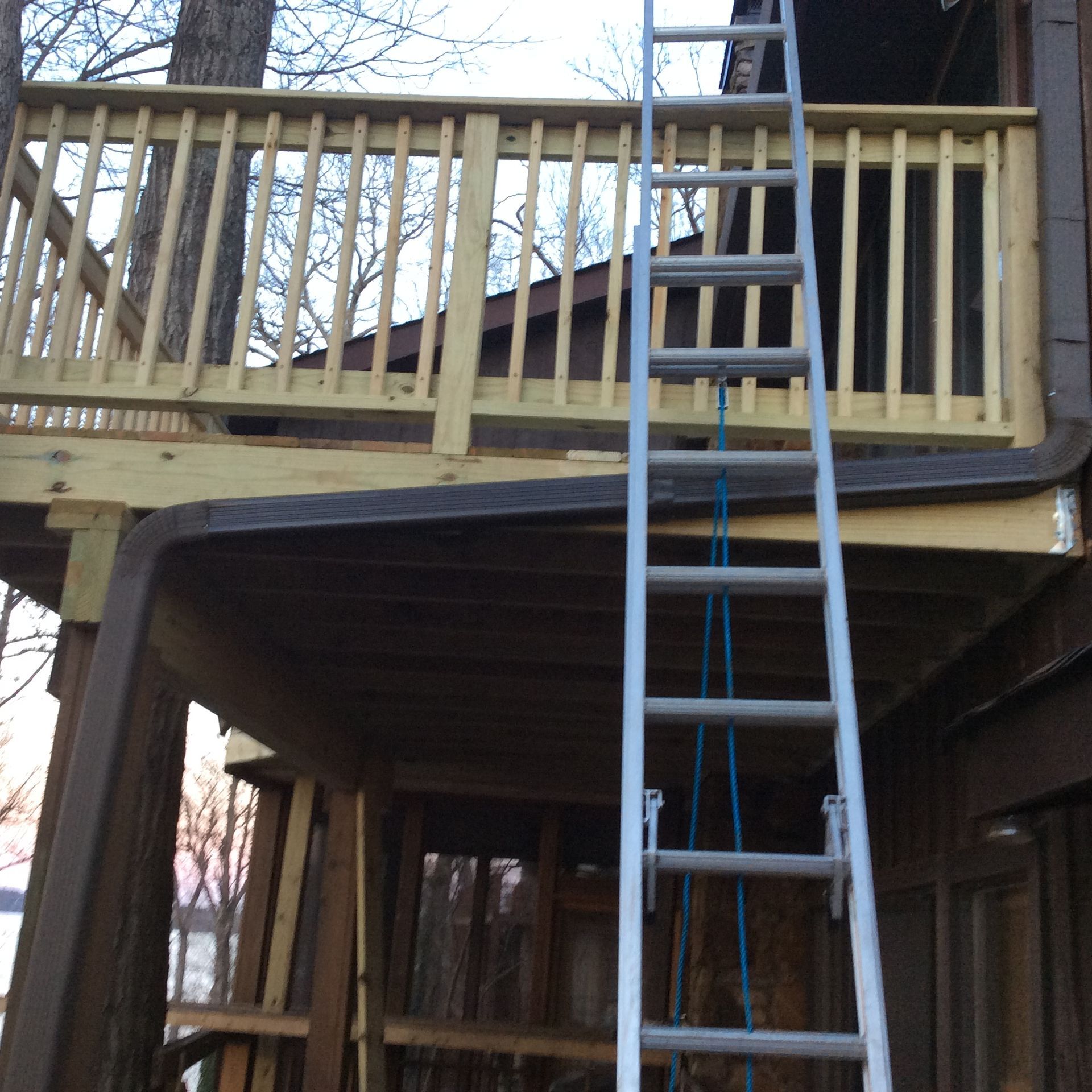 An extension ladder leans against a wooden deck under construction, showing the framework of a house in a wooded area.