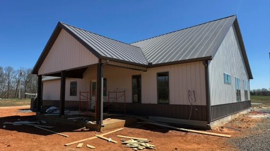 A modern house under construction featuring white vertical siding, a dark metal roof, and a covered porch on a dirt lot.