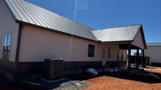 A side view of a house under construction with white siding, dark metal roofing, and a covered porch on a dirt lot.