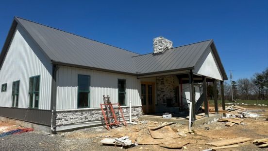 A partially constructed white farmhouse with a dark metal roof, stone accents, and a porch under a clear blue sky.