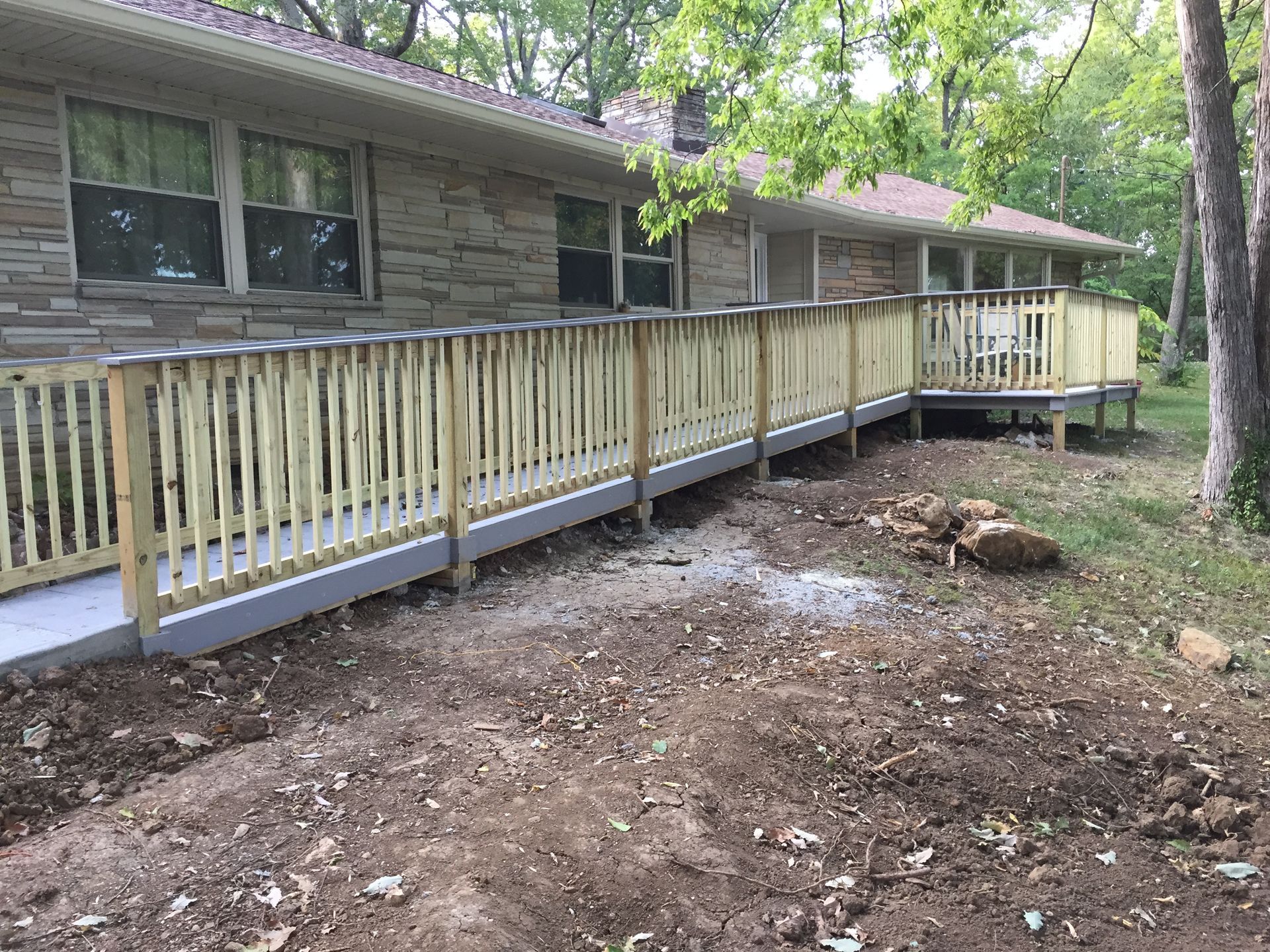 A long, wooden wheelchair ramp with railings installed along the side of a stone house in a wooded yard.