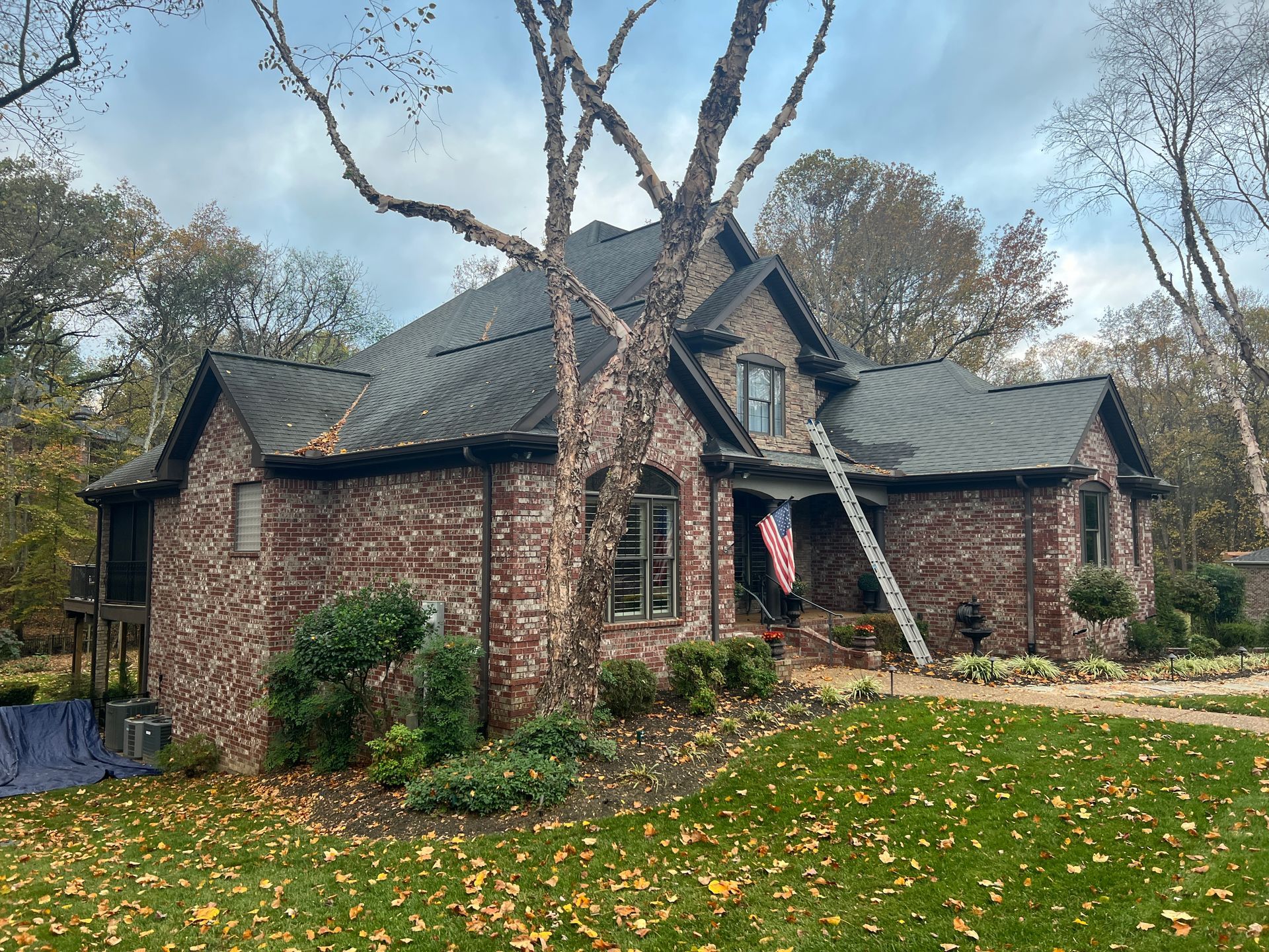 A two-story brick house with a dark shingled roof, a ladder against the side, and autumn leaves on the front lawn.