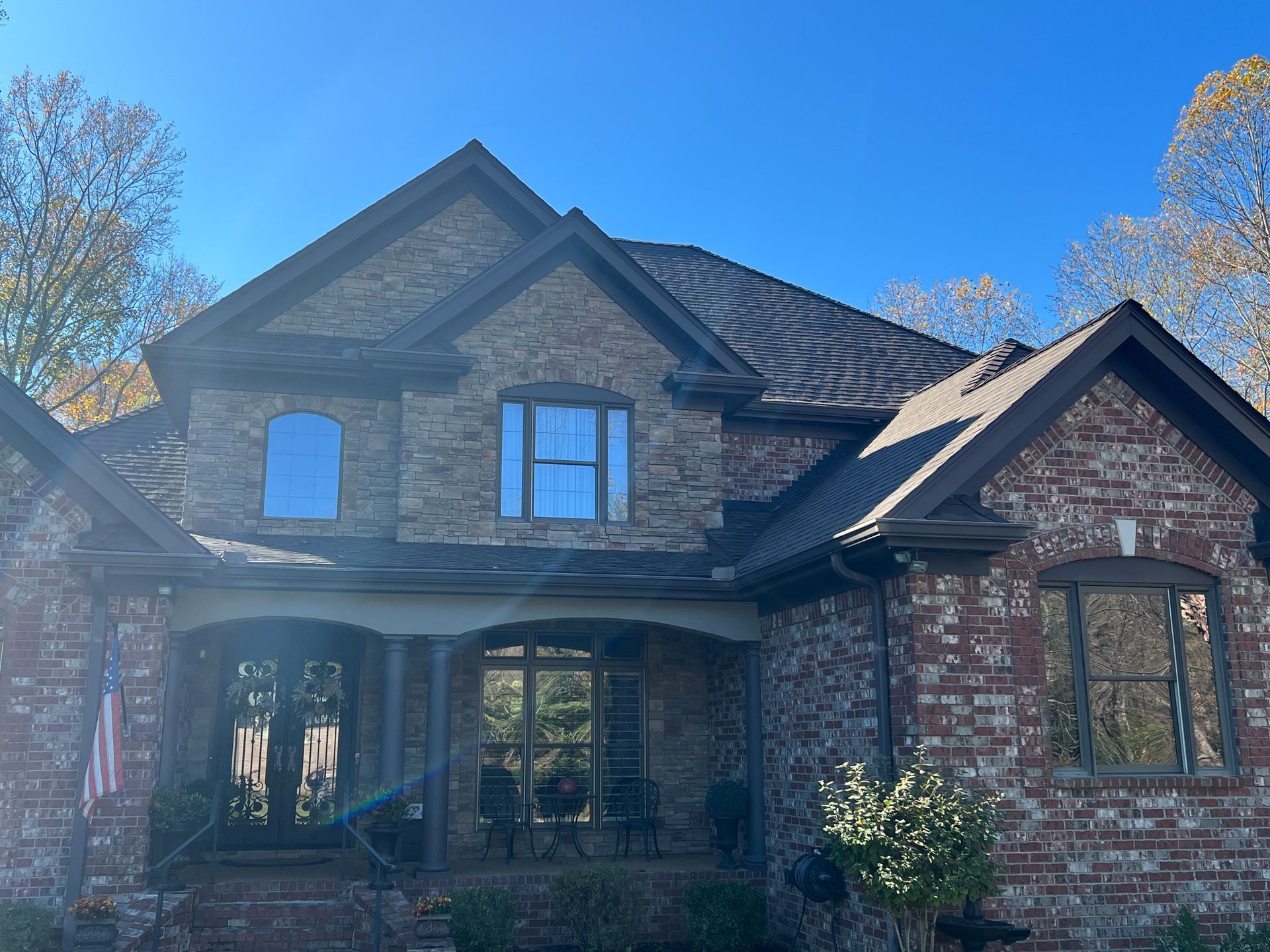 Two-story brick and stone house with a covered front porch, dark trim, and gabled roof under a clear blue sky.