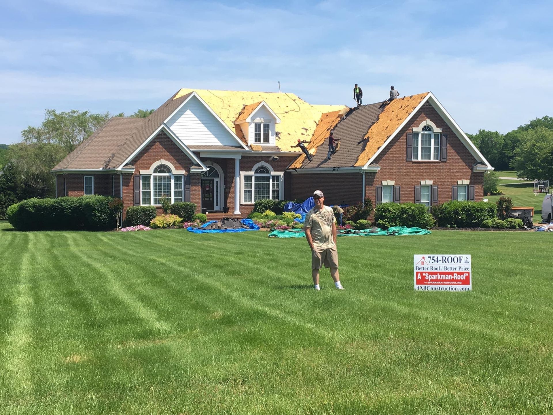 A person stands on a lawn in front of a brick house with a roof under construction and workers on the roof.