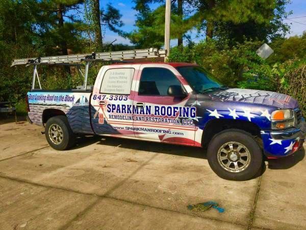 A white pickup truck with a roof ladder, wrapped in a patriotic American flag design and branded with Sparkman Roofing.