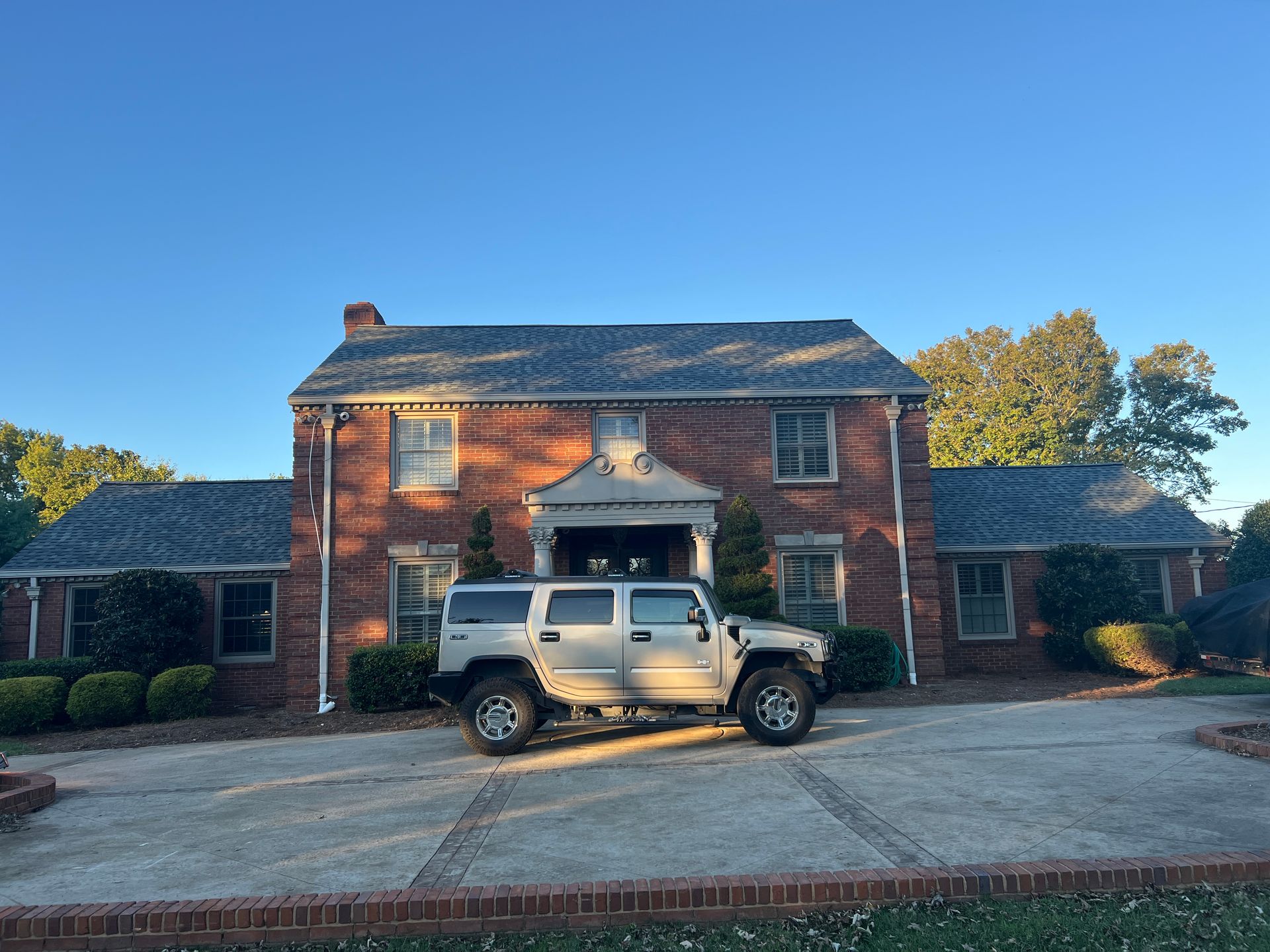 A light-colored Hummer SUV is parked in front of a two-story brick house with a gray shingled roof under a clear blue sky.