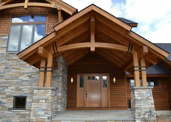 A front entrance of a modern home featuring a heavy timber truss porch, stone columns, and wood siding.
