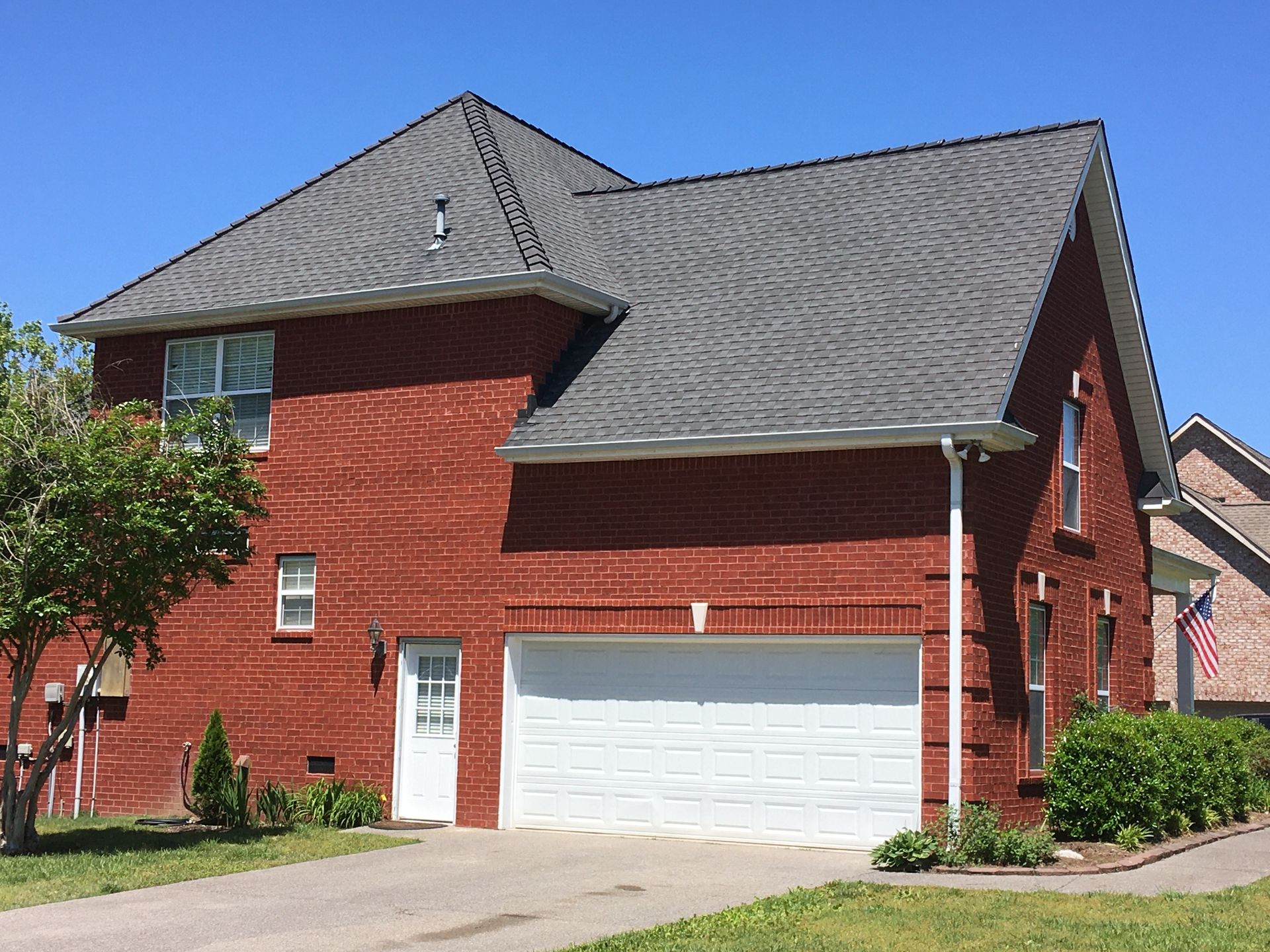 Two-story red brick house with a gray shingled roof, white garage door, and side entrance under a clear blue sky.