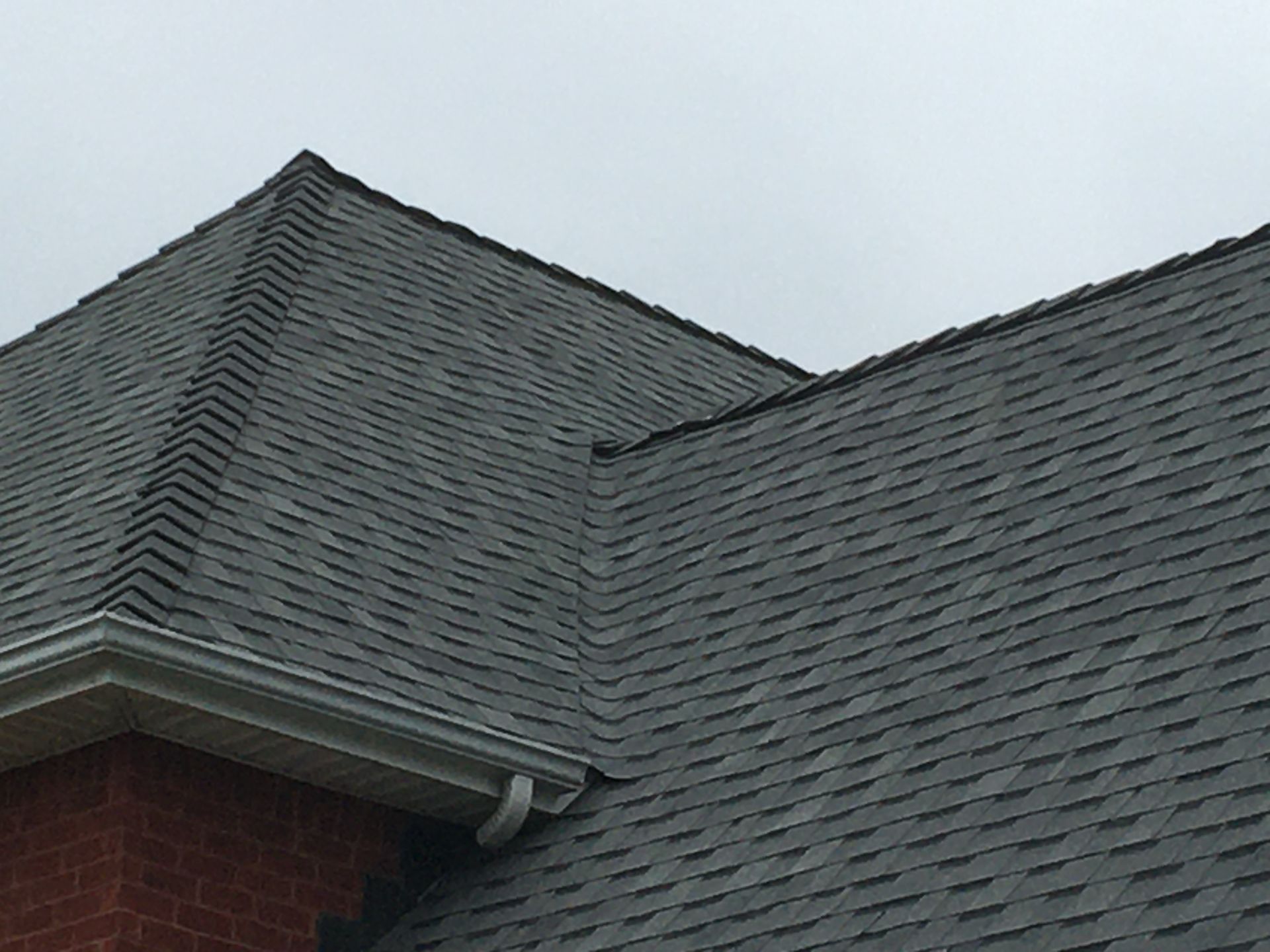 A close-up view of the corner of a dark grey asphalt shingle roof on a red brick building against an overcast sky.