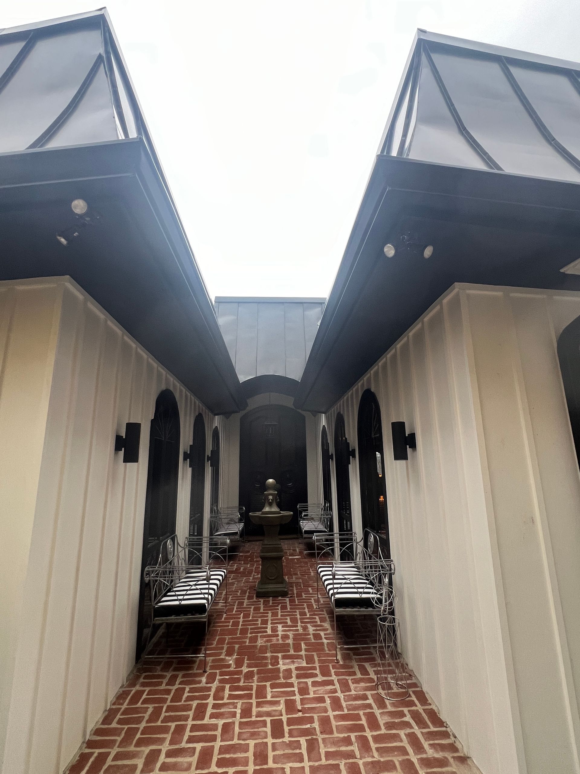 A brick-paved courtyard between two light-colored buildings, featuring a central fountain and dark iron benches.