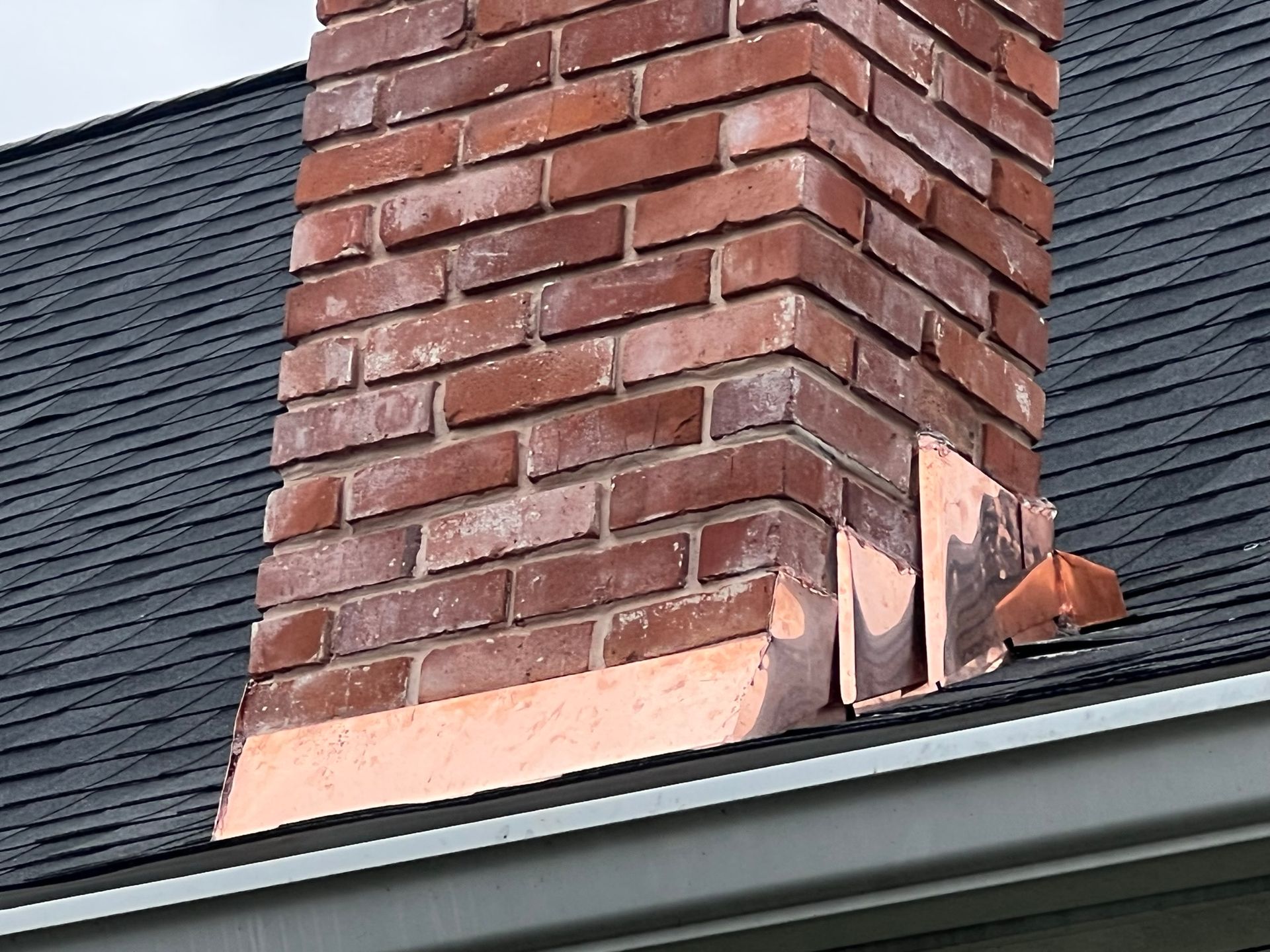 A brick chimney rising from a shingled roof with copper flashing partially detached at the base.