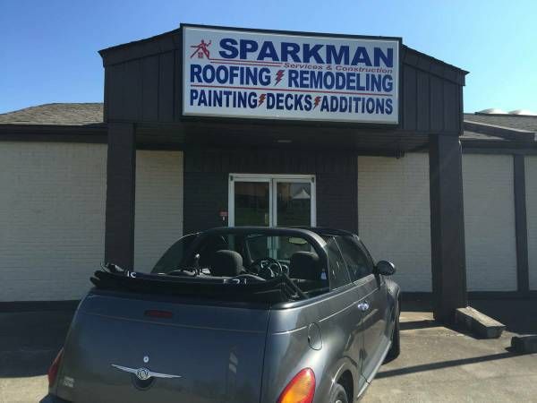 A grey convertible parked in front of a Sparkman construction company building featuring signs for roofing and remodeling.