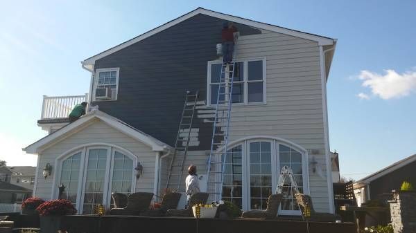 Two painters on ladders work on repainting the beige siding of a house to dark blue.