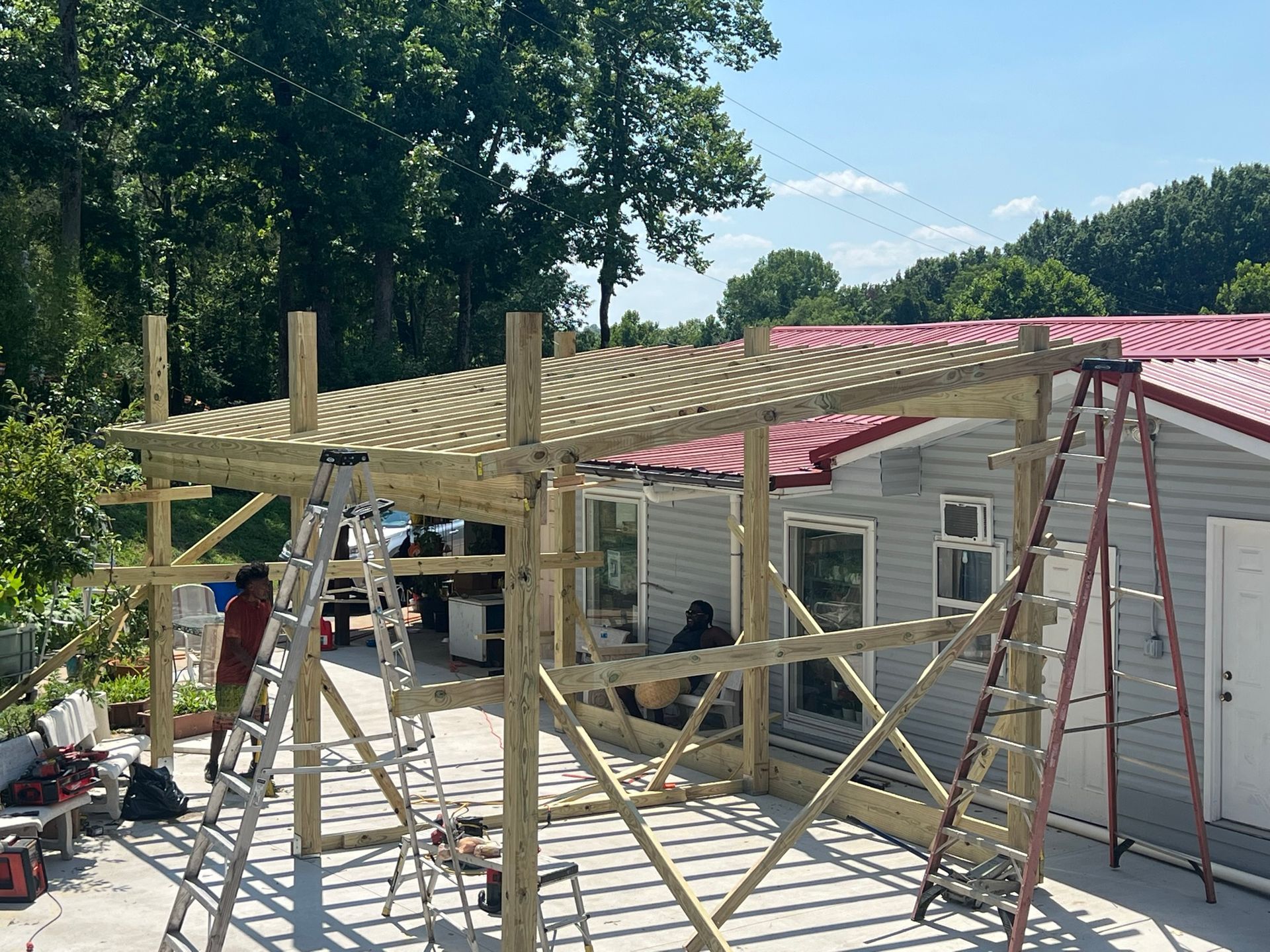 A partially constructed wooden patio cover is being built attached to a house with gray siding and a red metal roof.