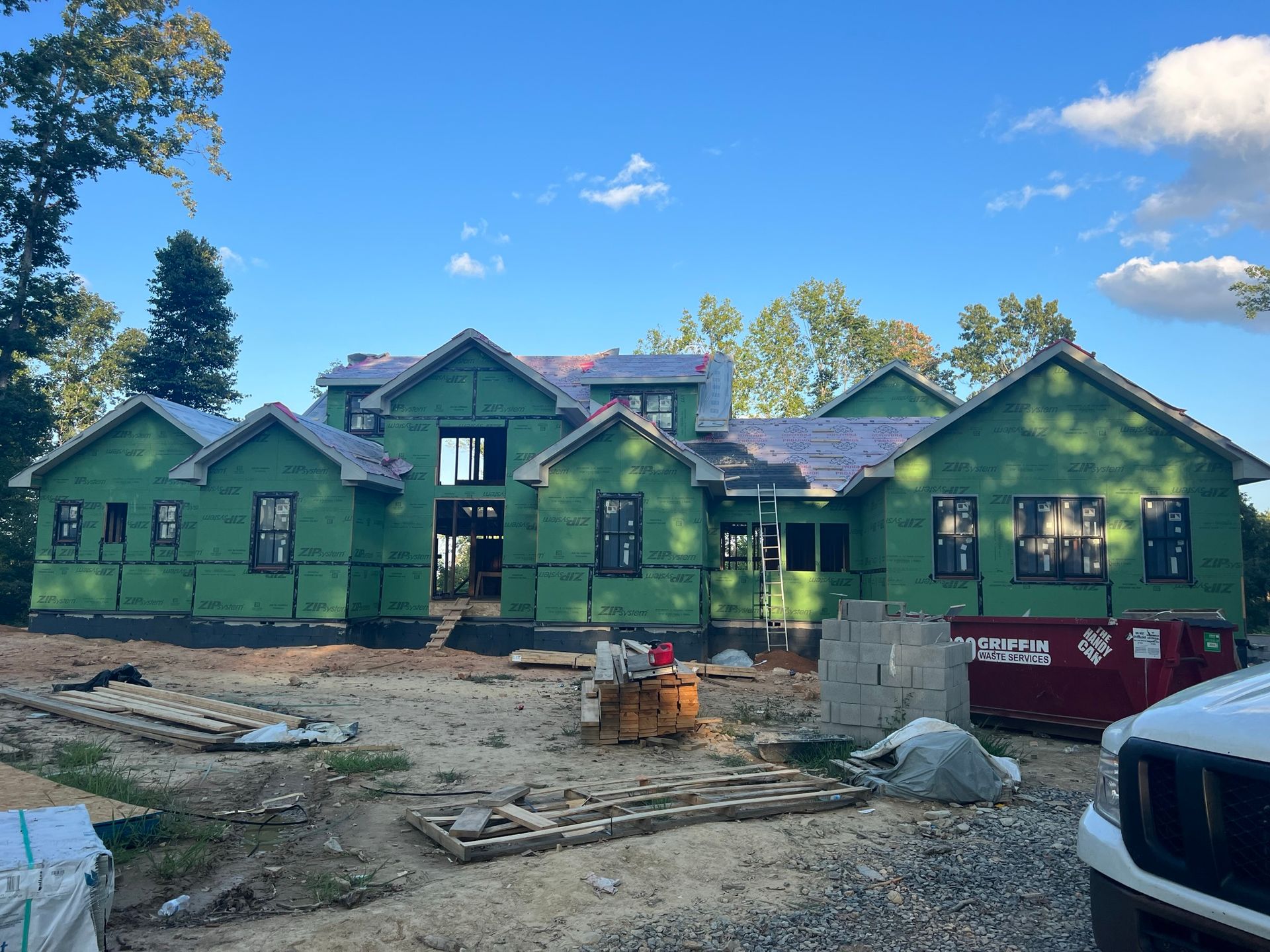 A large, green-sheathed house under construction, featuring multiple gables and installed windows on a sunny day.