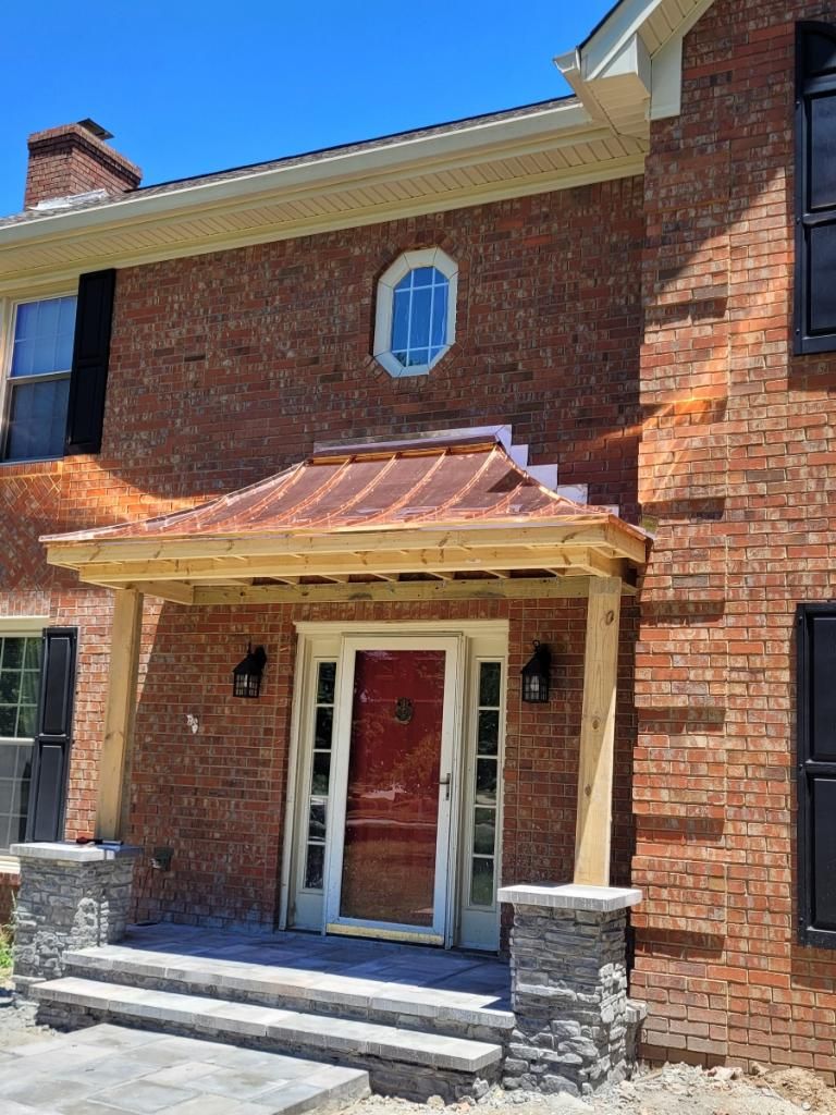 A brick house front entrance featuring a new copper-roofed porch, wooden support beams, and stone-base pedestals.