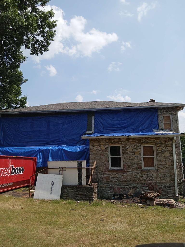 A stone house under renovation with blue tarps covering parts of the roof and siding, next to a red dumpster.