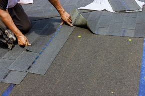 A worker installs dark gray asphalt roof shingles onto a residential roof surface.