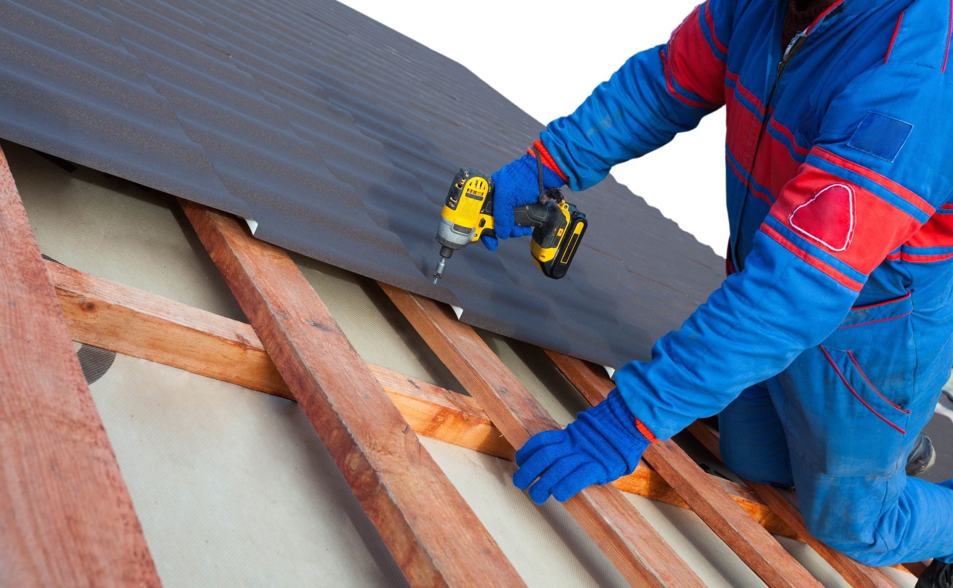 A construction worker in a blue uniform uses a power drill to fasten a black roof panel to wooden beams.
