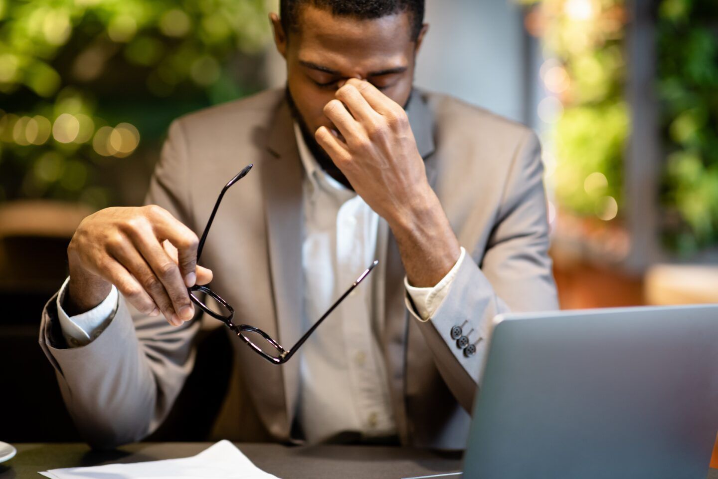 Man in suit stressed, rubbing eyes, holding glasses near laptop.