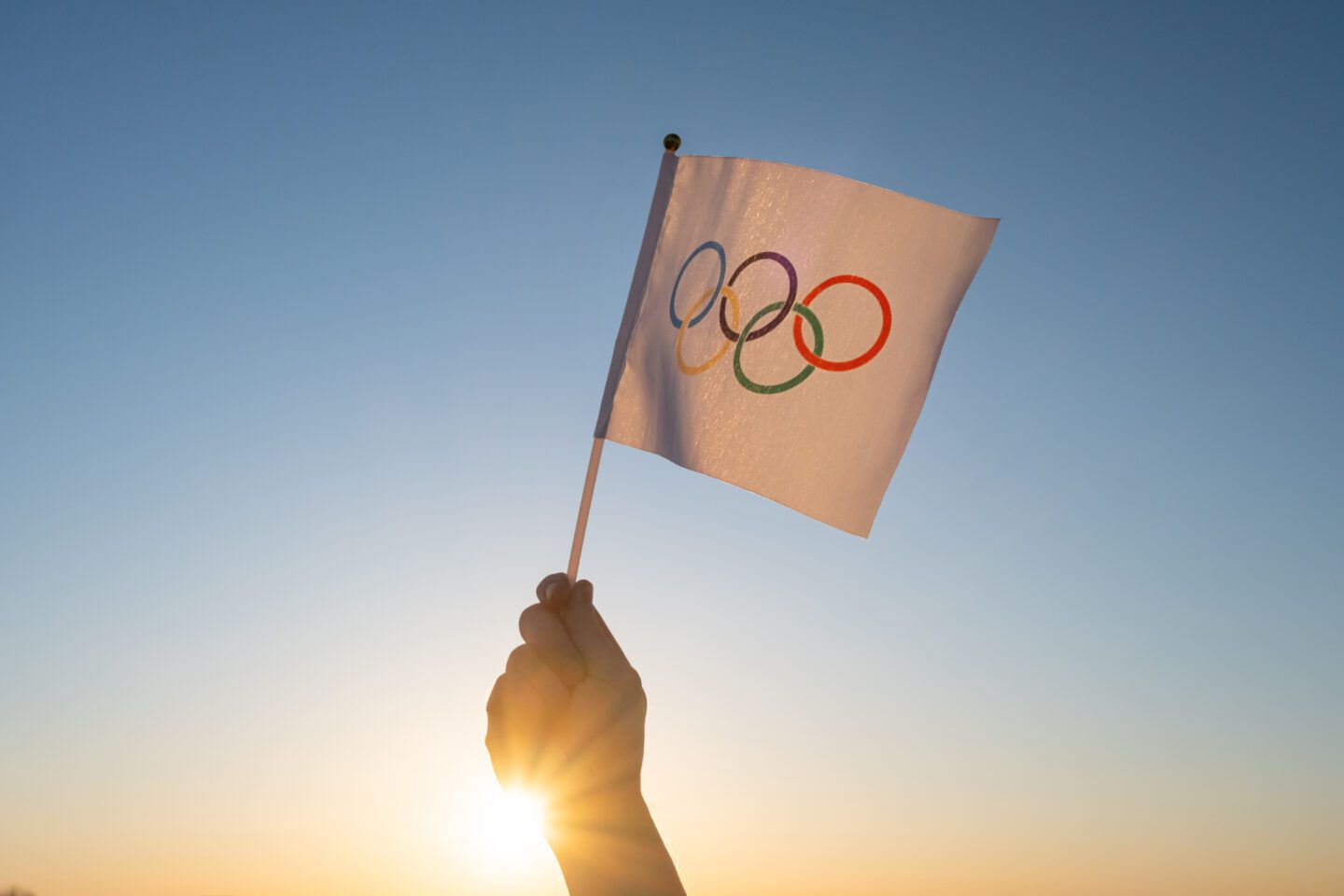 Hand holding Olympic flag against a bright sunrise. Rings: blue, yellow, black, green, red.