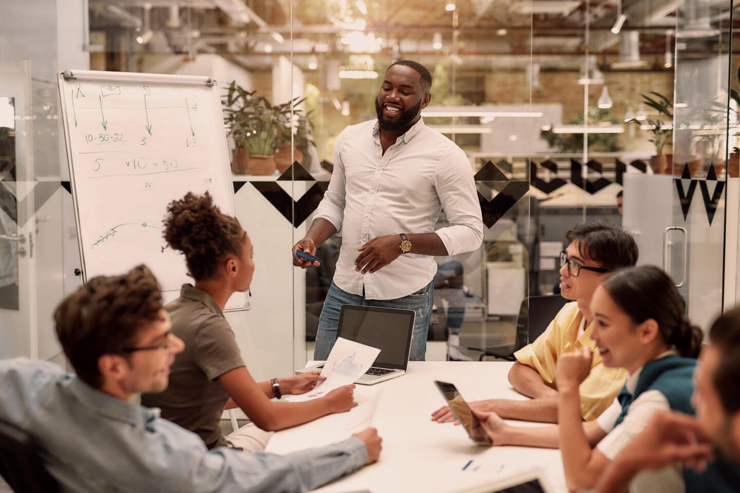 Man presents to team at table in modern office; whiteboard, papers, and devices are present.