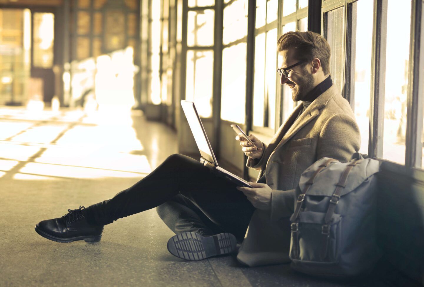 Man sitting on floor, working on laptop and phone, smiling. Suit jacket, black pants, and backpack.