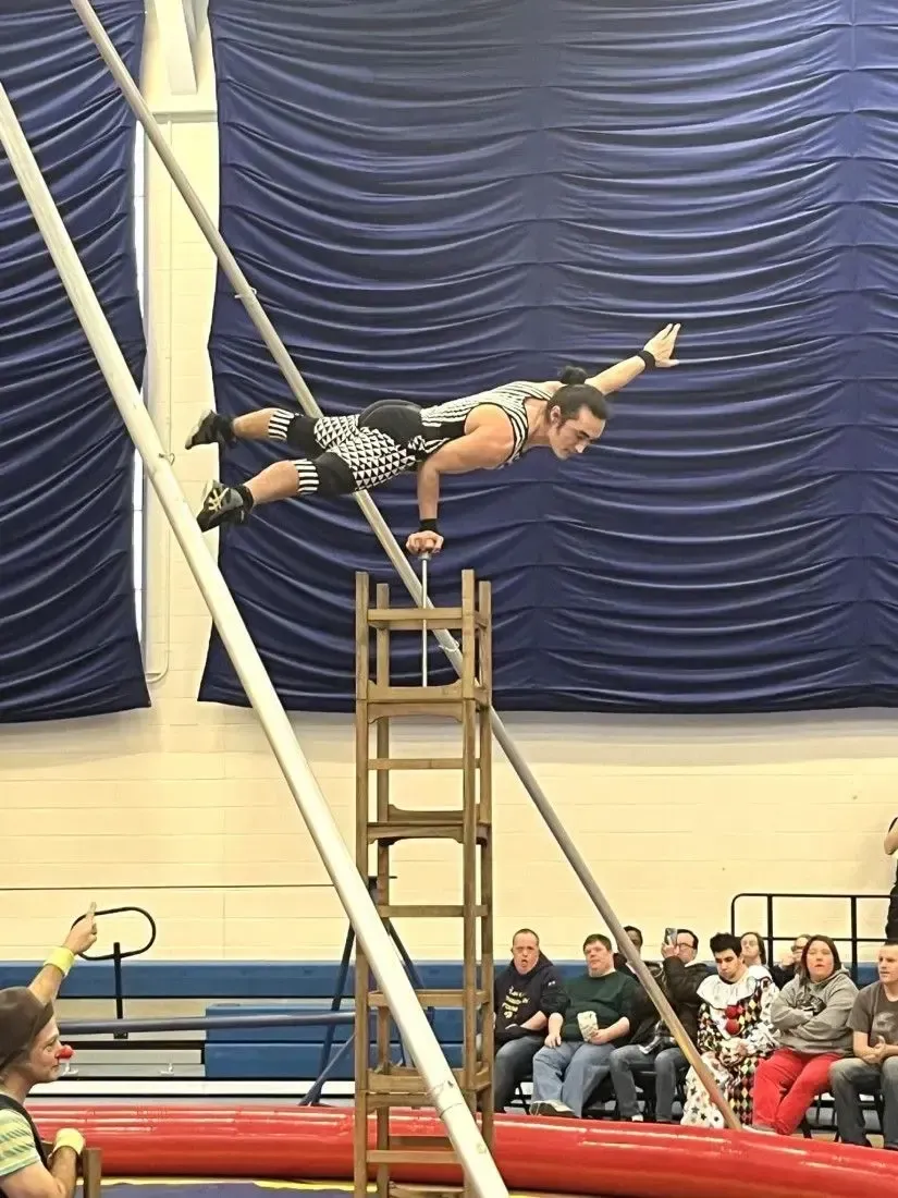 Acrobat balances on a ladder, arms outstretched, performing a stunt. Audience watches in a gymnasium.