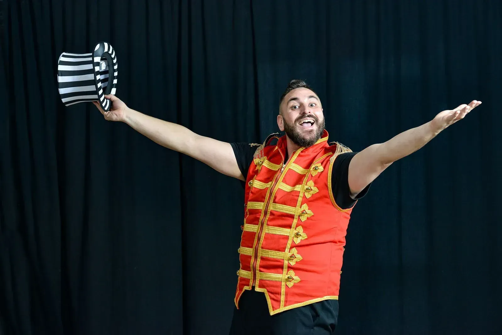 A man in a circus costume is holding a top hat with his arms outstretched.