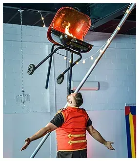 A man is balancing a wheelbarrow on a pole with fire coming out of it.