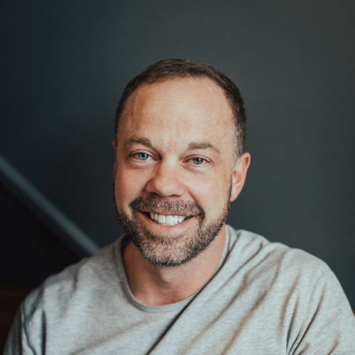 Smiling man with short brown hair and a beard, wearing a gray t-shirt.