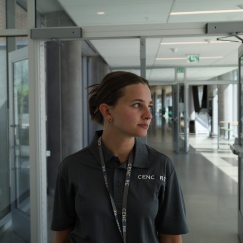 Woman in grey polo shirt and lanyard stands in a brightly lit hallway, looking right.