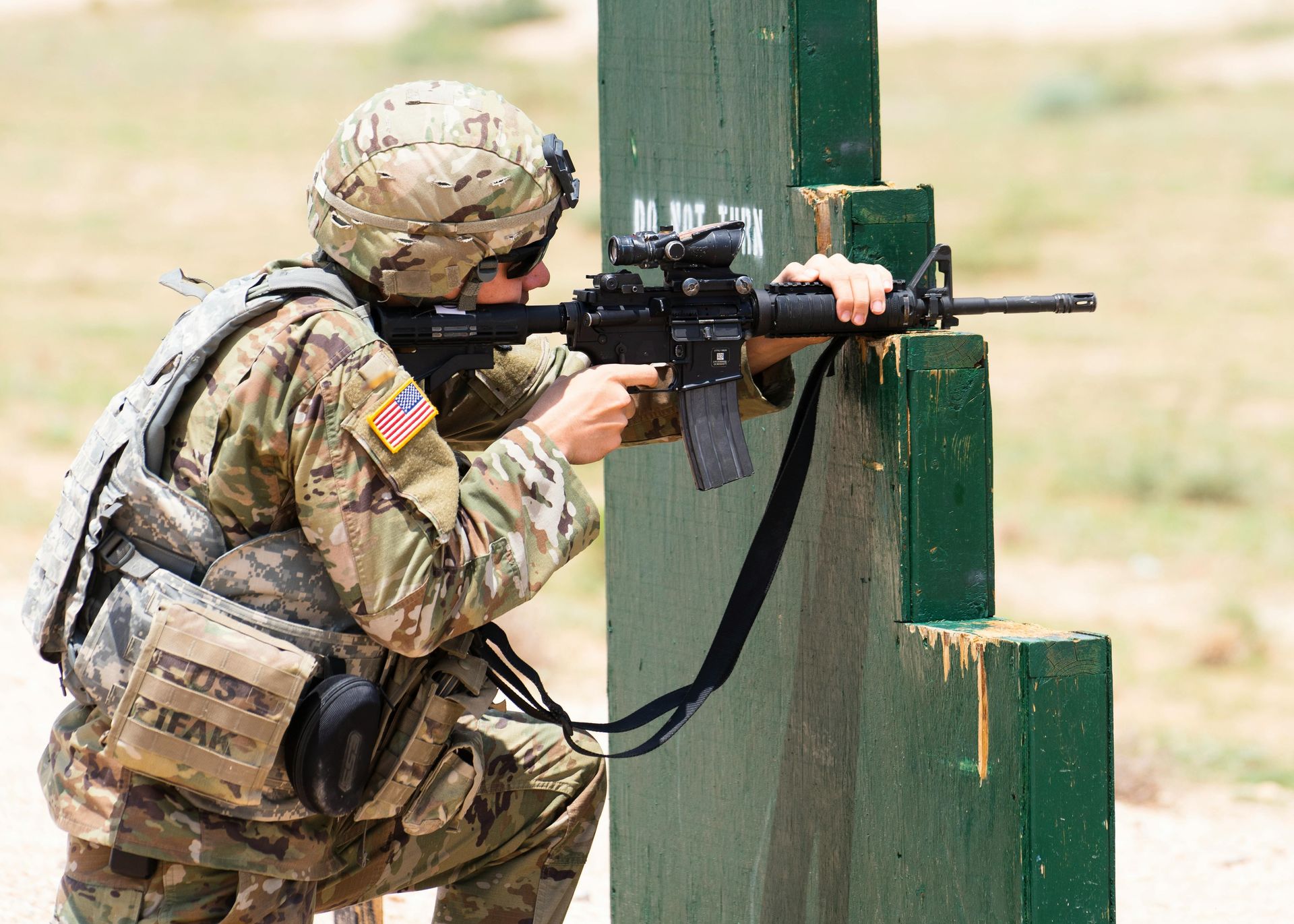 Soldier aims rifle at a range, kneeling behind a green barrier.