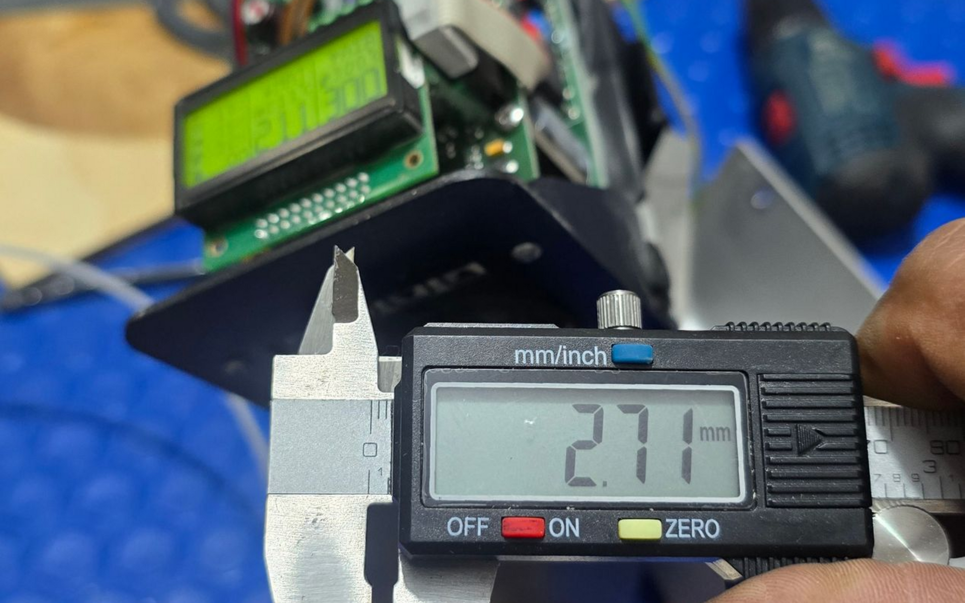 A person measures a black metal component with digital calipers, reading 2.71 mm. An electronic device is in the background.