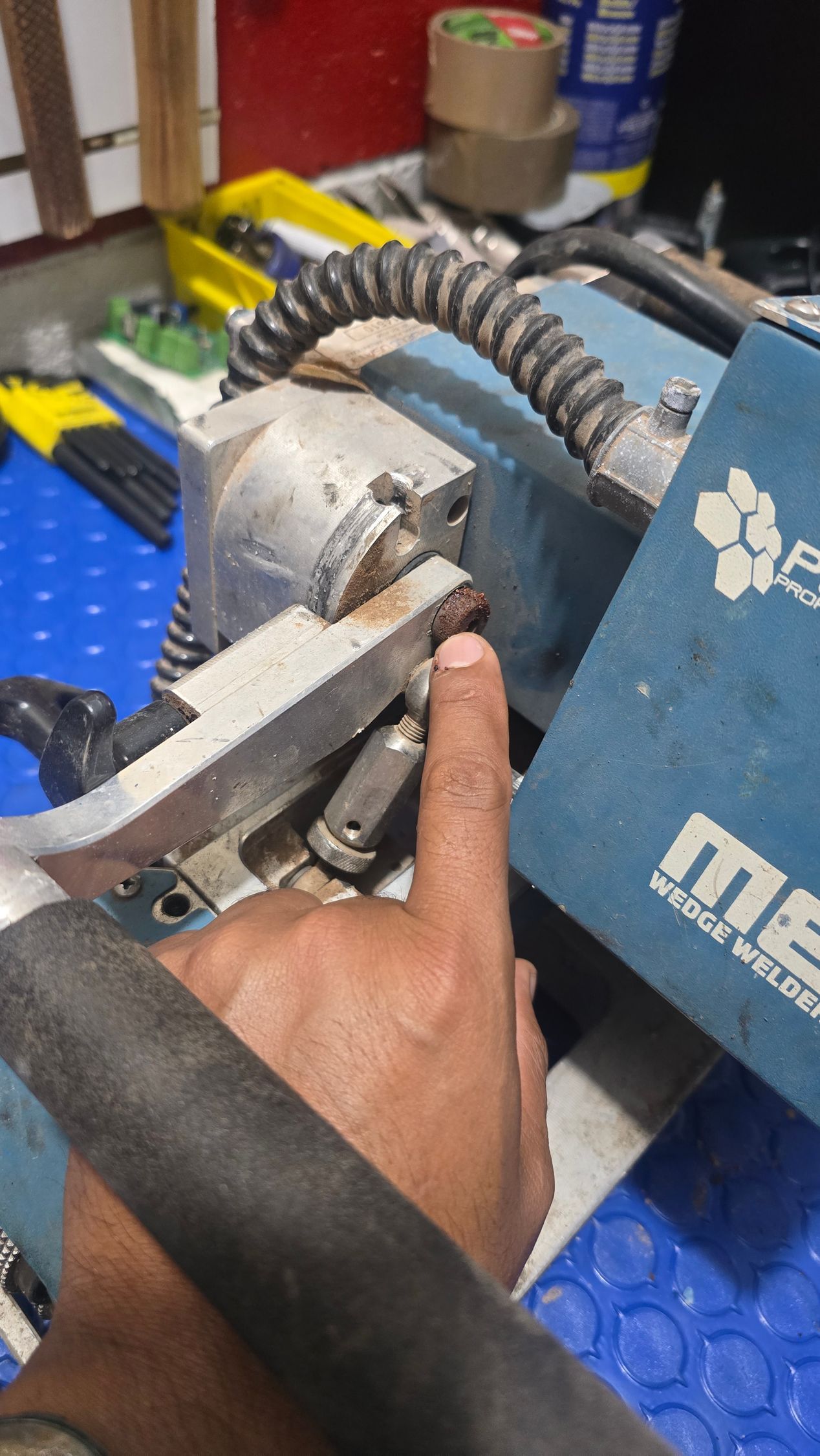 Person's finger pointing at a metal welding machine part, possibly a nozzle. Blue machine with a black handle.