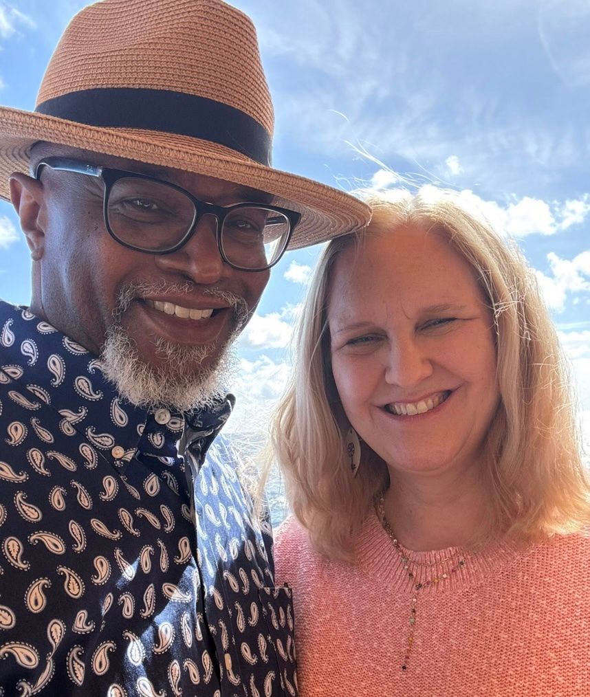 Two people smiling, outdoors. Man wearing hat and glasses, patterned shirt. Woman in pink sweater. Blue sky background.