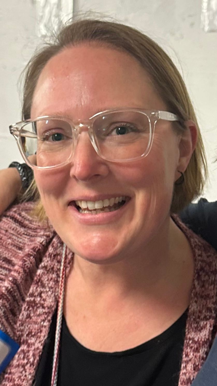 Woman smiling, wearing clear-framed glasses and a red cardigan, looking towards the camera.