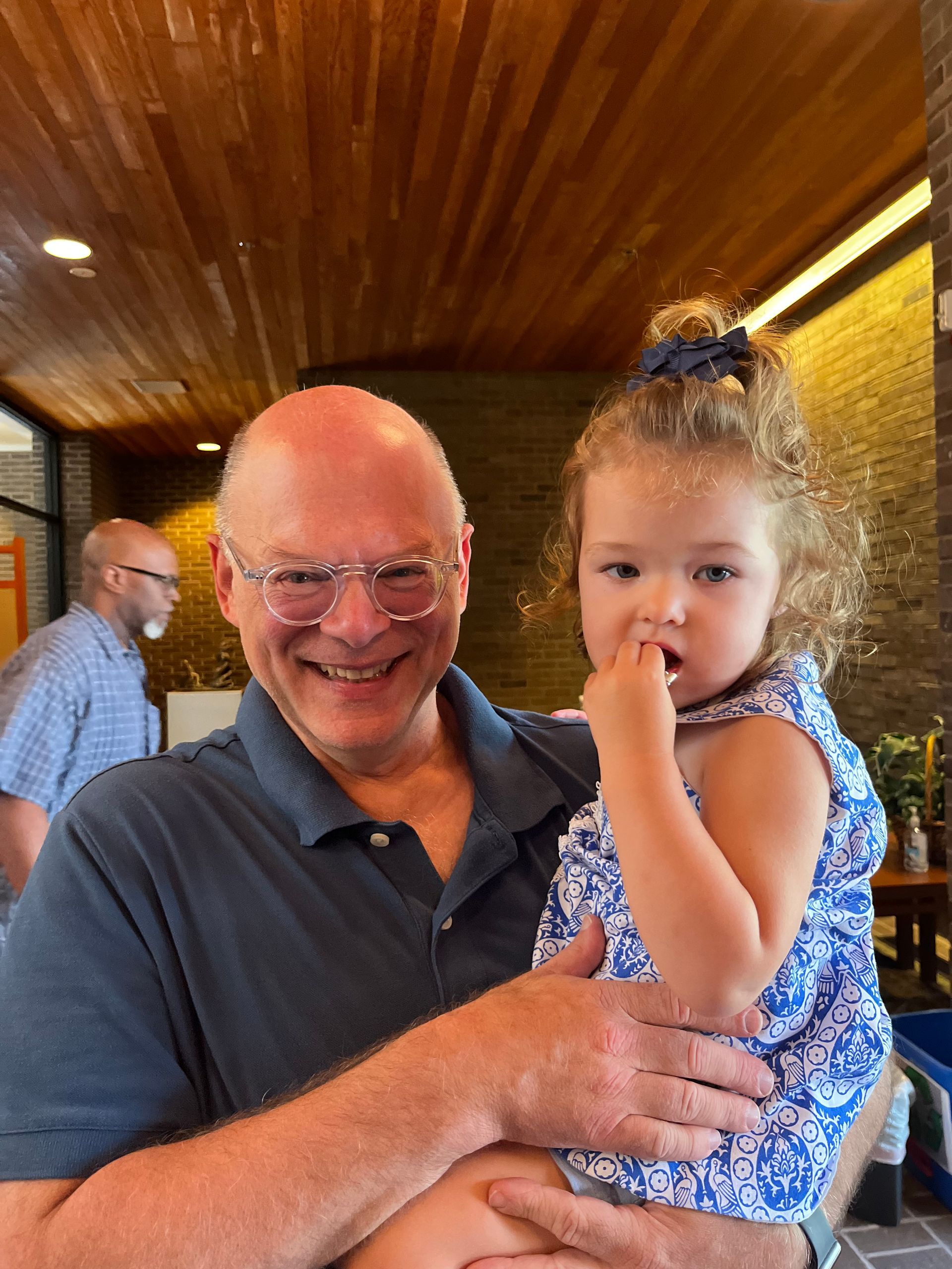 Man holding a child, both smiling indoors. Man wears glasses and a blue shirt. Child has curly hair and a blue patterned dress.