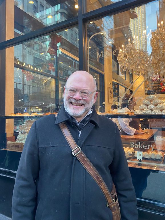 Man with glasses smiles in front of a bakery window, wearing a black coat and cross-body bag.