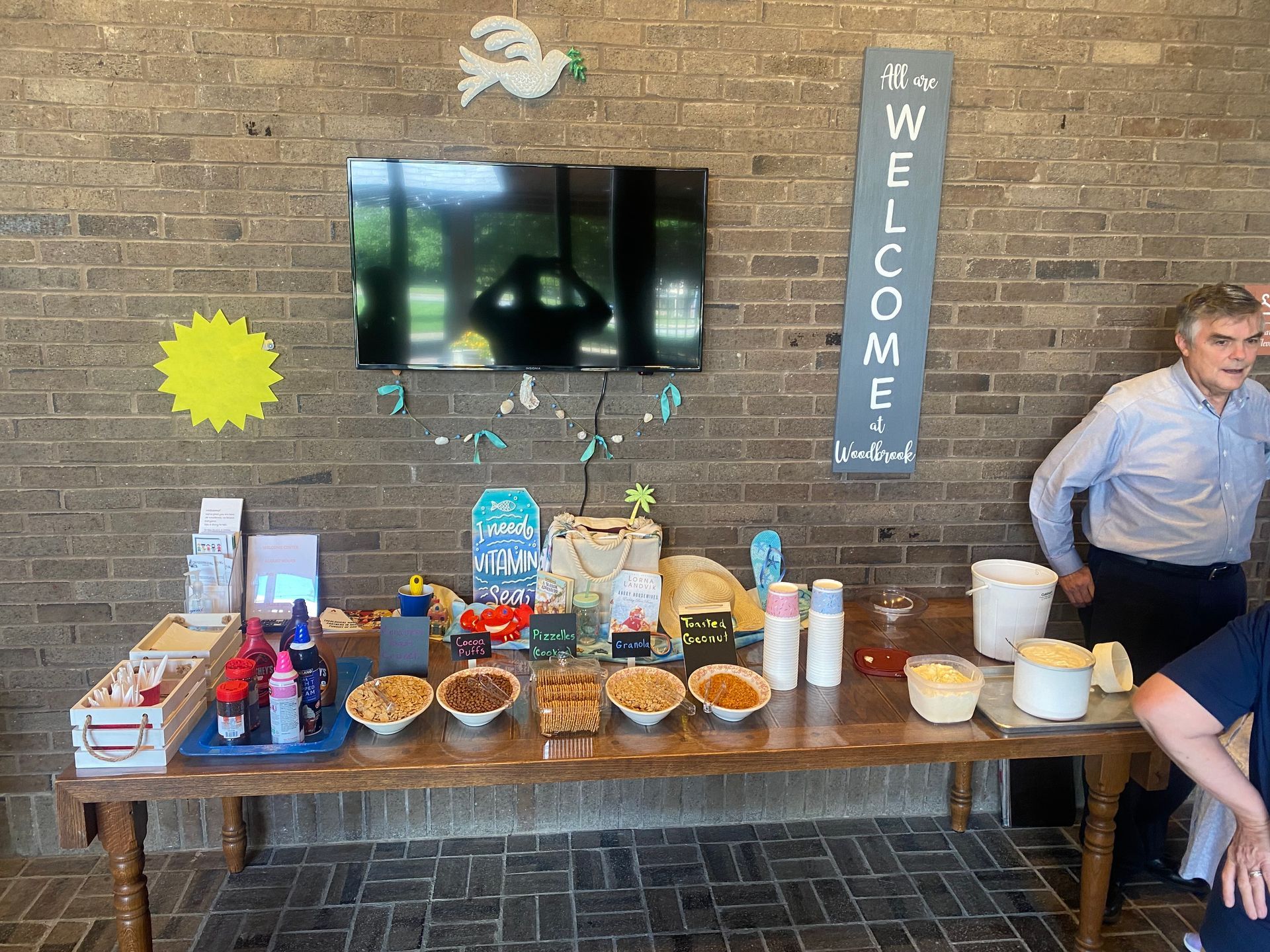 Table of snacks, drinks, and welcome sign against a brick wall. 