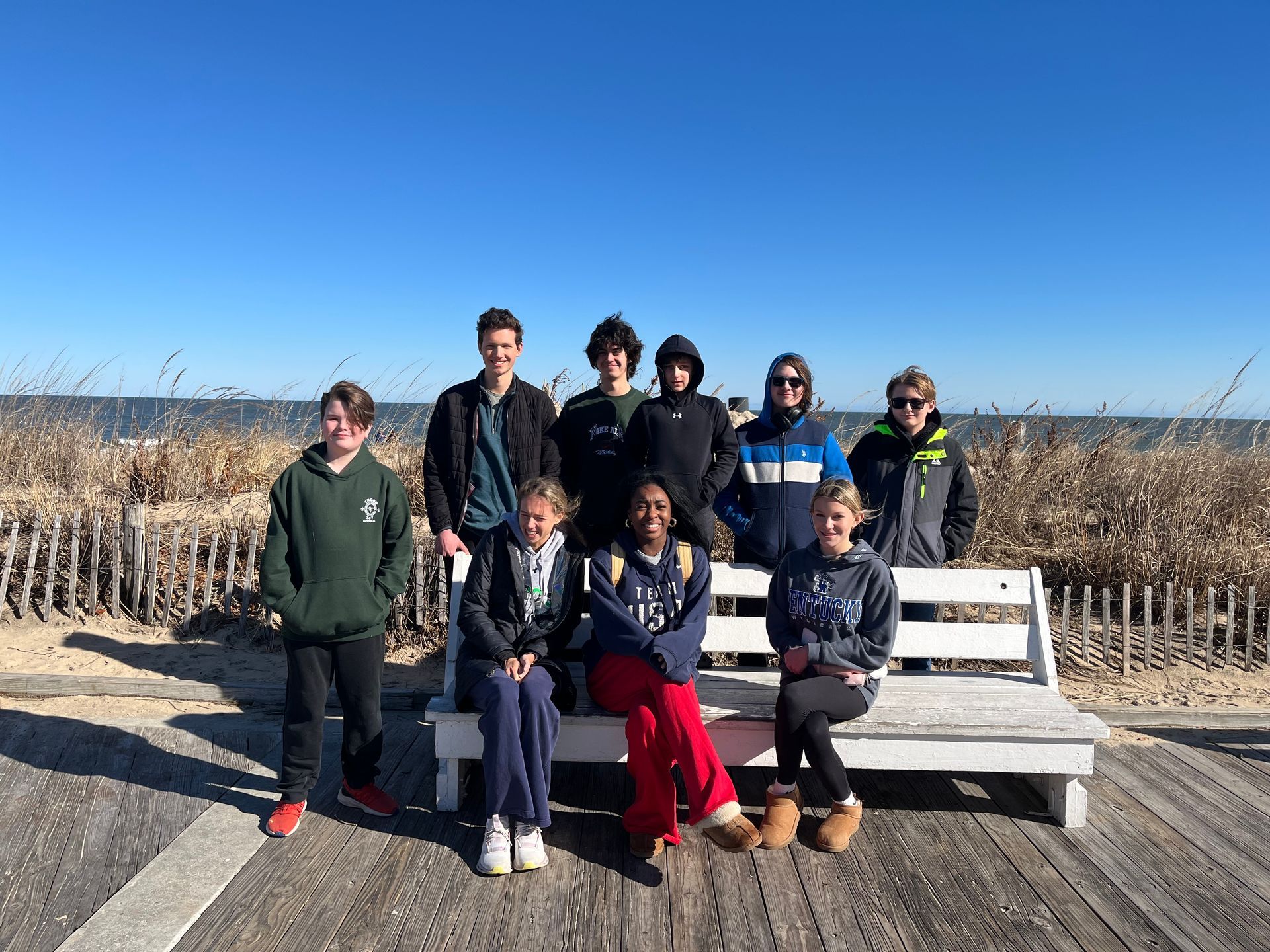Youth group on a boardwalk bench by a beach. Clear blue sky.