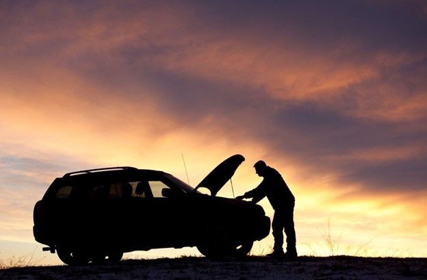 A man is looking under the hood of a car at sunset.