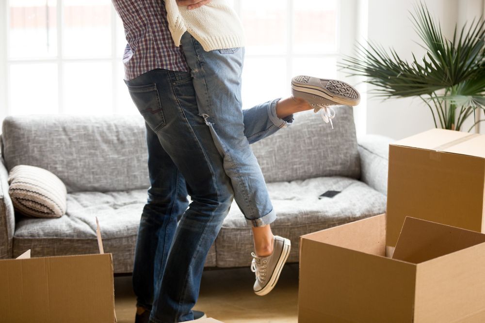 Couple celebrating move, man lifting woman, boxes, sofa, sunny room — Go With Joe REMOVALS in Rockhampton, QLD