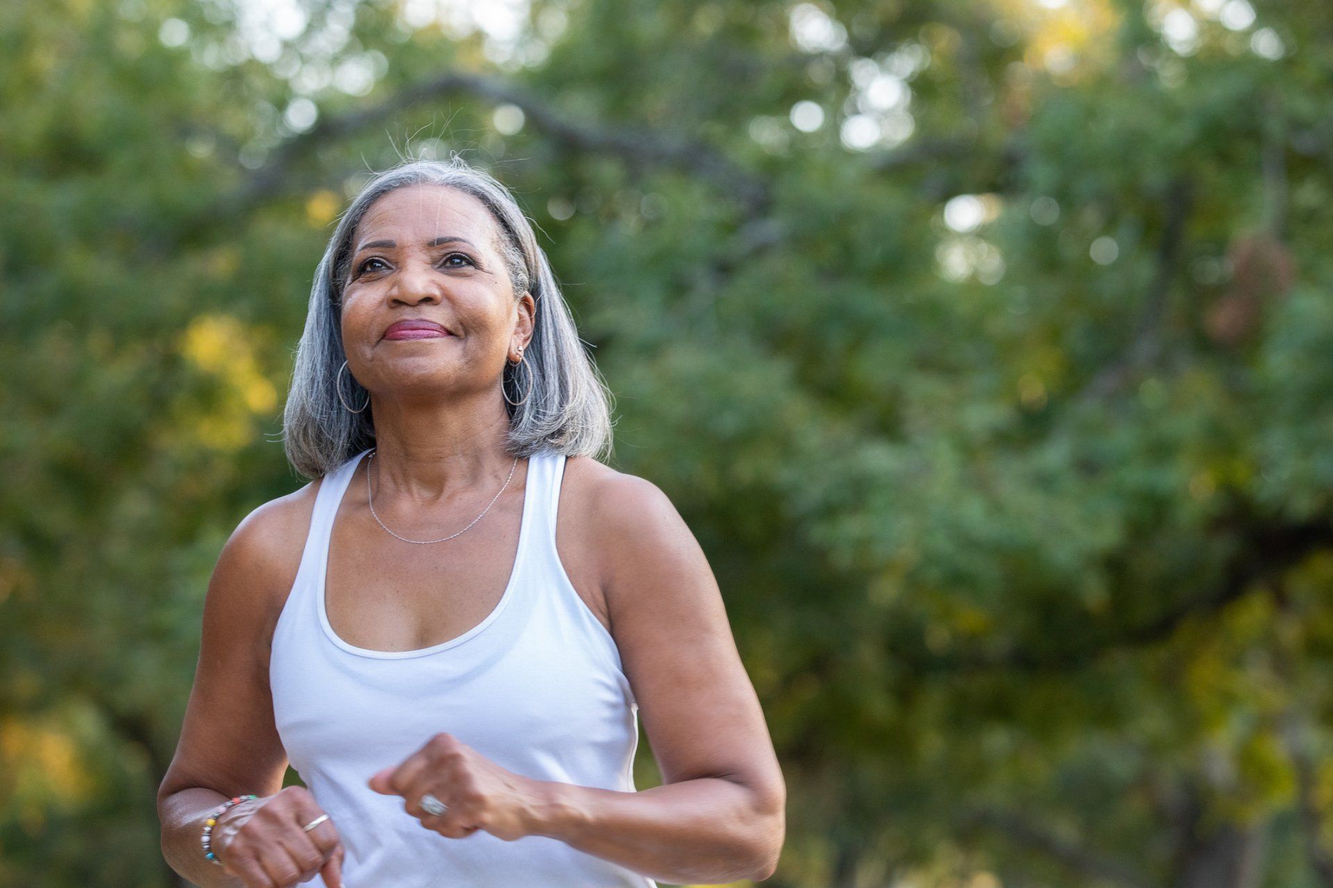 Woman in a white tank top jogging outdoors, smiling, with a backdrop of green trees.