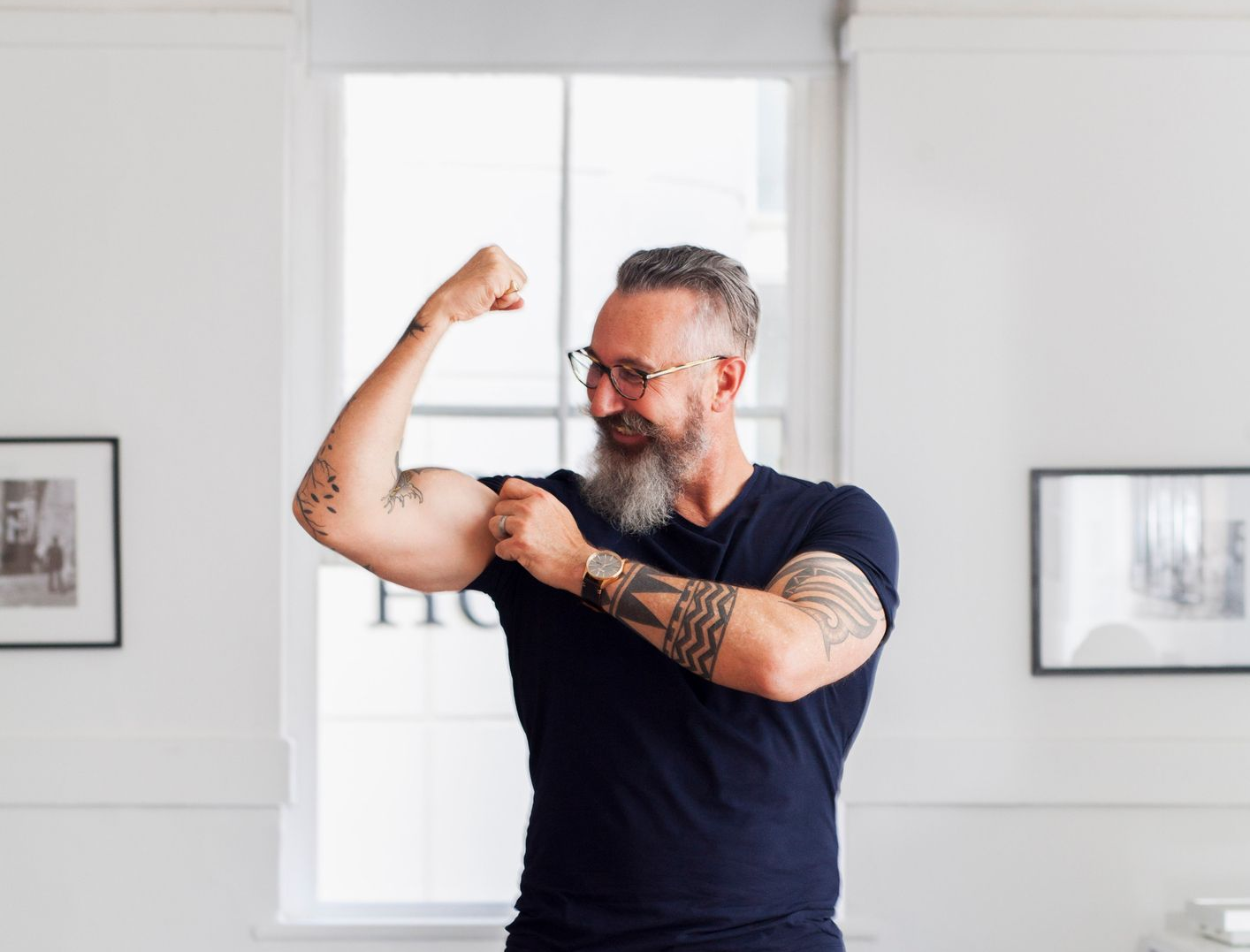Man flexing arm, showing off bicep. Tattoos on arms. Wearing glasses, watch, and dark shirt. Indoors, near a window. Man flexing arm, showing off bicep. Tattoos on arms. Wearing glasses, watch, and dark shirt. Indoors, near a window.