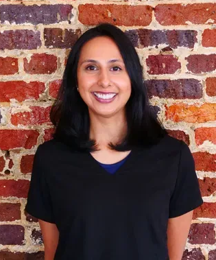 Doctor wearing glasses, stethoscope, and lab coat, holding clipboard, talking on screen.