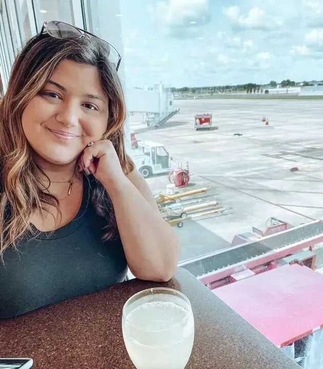 Woman and man in suits, looking at laptop and tablet on wooden table, discussing outdoors. Woman at airport, smiling, with drink. Window view of tarmac and jet bridge.